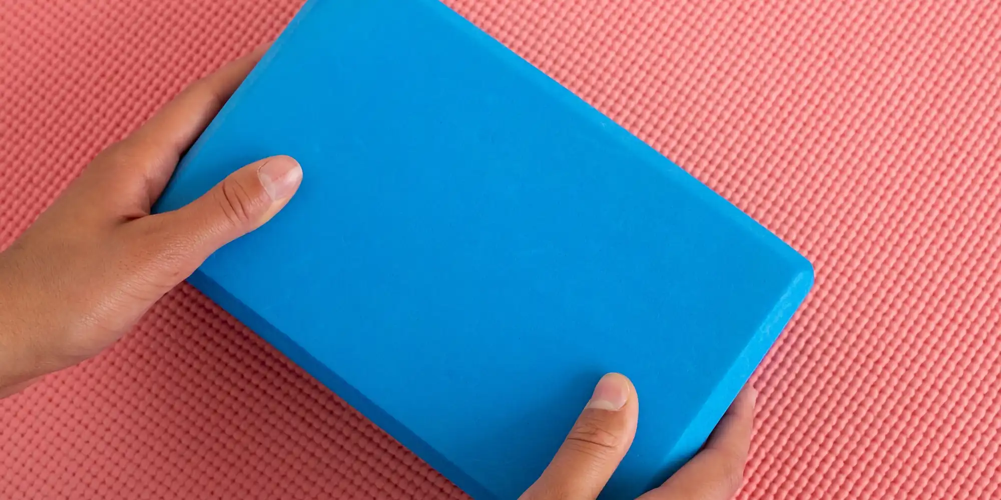 A pair of hands gently holding a blue yoga block, positioned on a textured pink yoga mat.