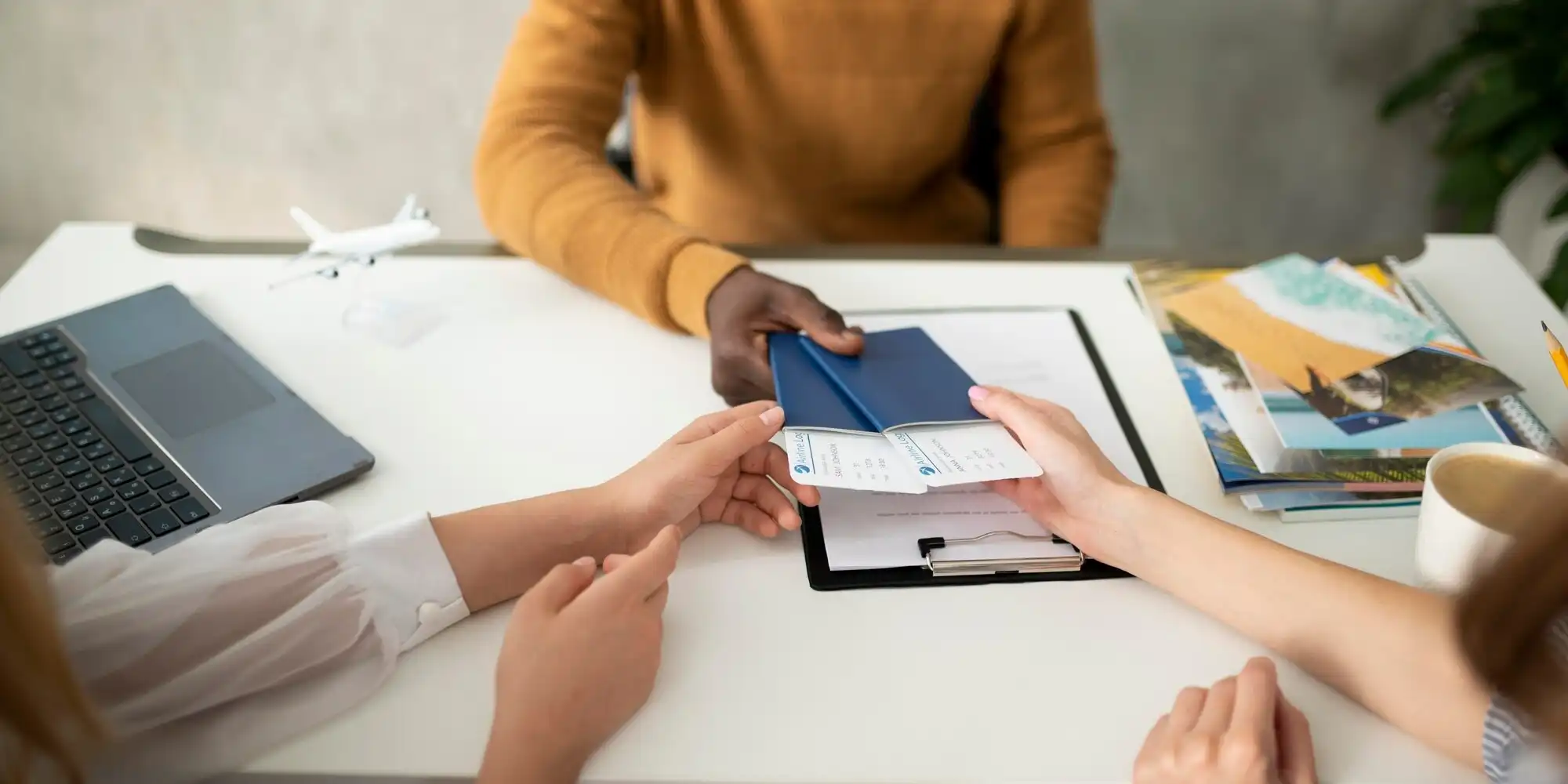There are two people sitting at a table. One of them was wearing a yellow top, only part of whose body could be seen. A laptop, a folder and some magazines were on the table. There is also a white model drone sitting on the table. Two people are exchanging a few cards. One of them is holding a few cards with words and logos while the other is holding out his hand to receive them. There is also a cup on the table. Overall, this photograph presents a professional work scene where two people are engaged in some sort of exchange or trading activity.