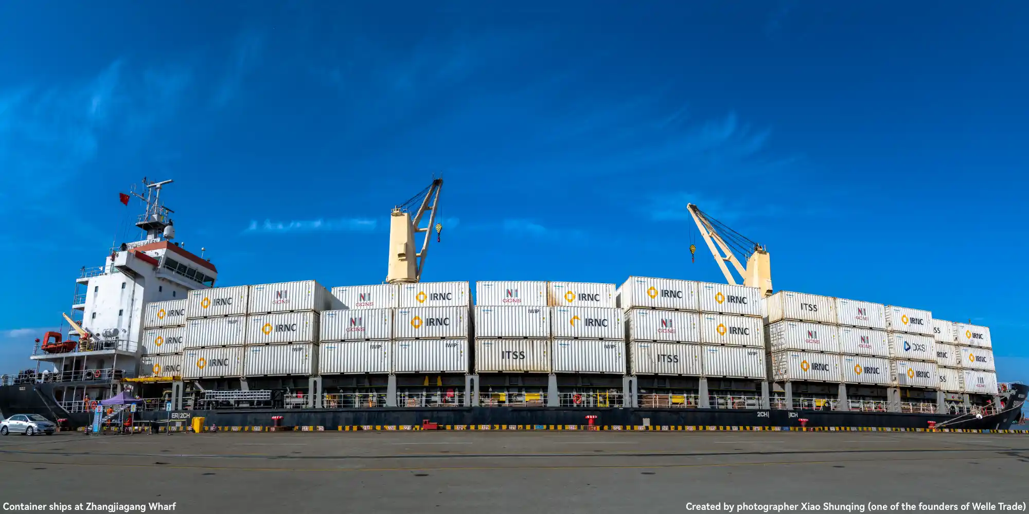 Container ship at Zhangjiagang Wharf, loaded with white containers marked with "IRNC" and "ITSS", equipped with ship cranes, under a clear blue sky; this photo is created by Xiao Shunqing (one of the founders of Welle Trade).