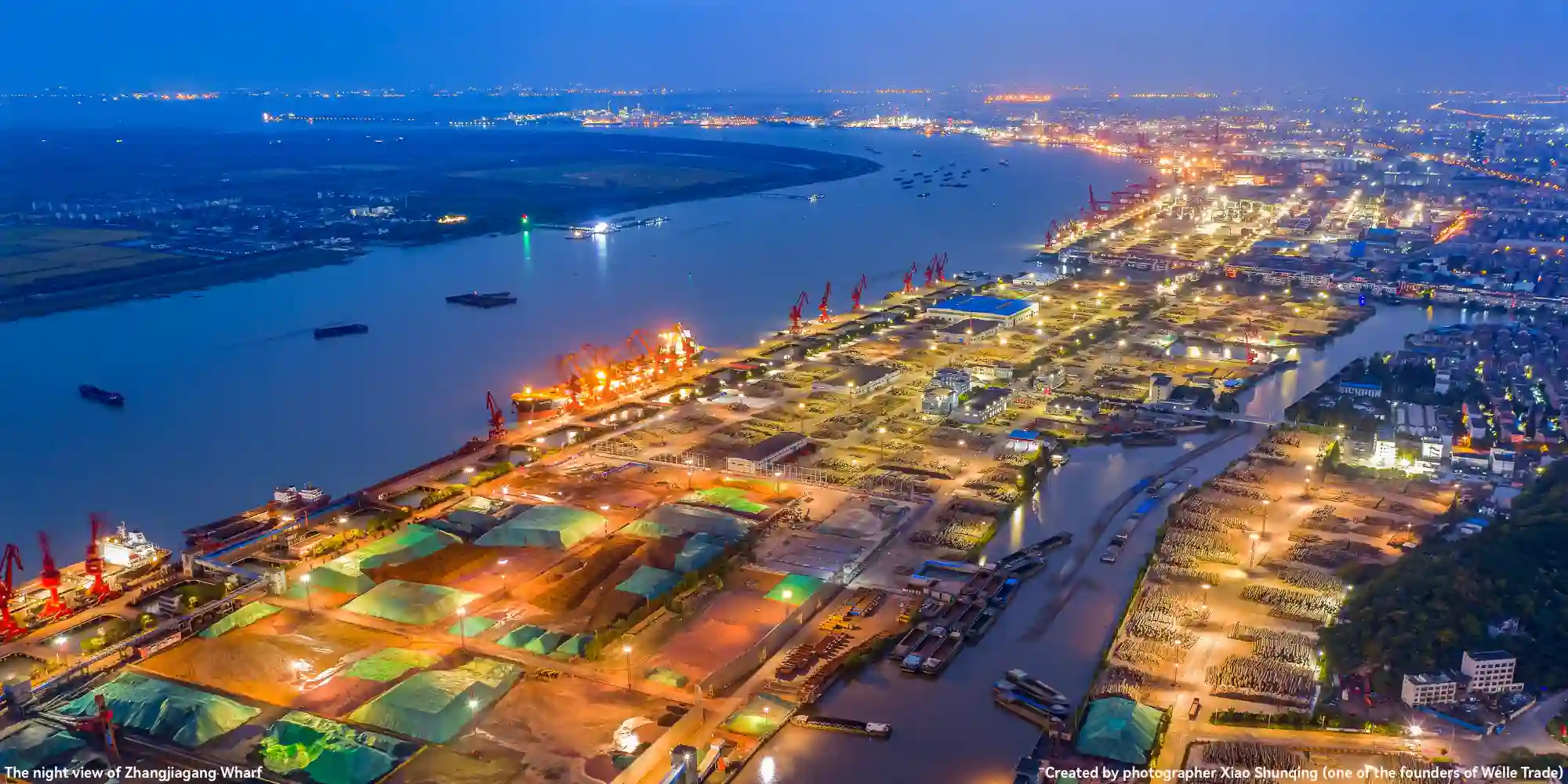 Aerial night view of Zhangjiagang Wharf: featuring a wide river with sailing ships, illuminated port facilities (including red cranes), and distant city lights; this shot is captured by Xiao Shunqing (one of the founders of Welle Trade).