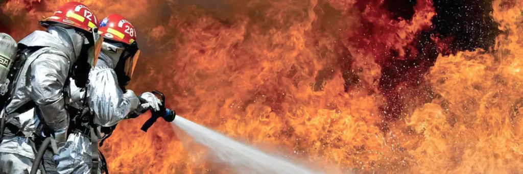 Two firefighters in silver heat-resistant suits and red helmets (marked with numbers) spraying a powerful jet of water from a hose to combat large, raging orange and red flames in the background.